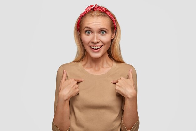 Wondered lucky female with surprised cheerful expression points at herself, asks whether she is really winner, cant believe in success, wears red headband and casual beige sweater, isolated