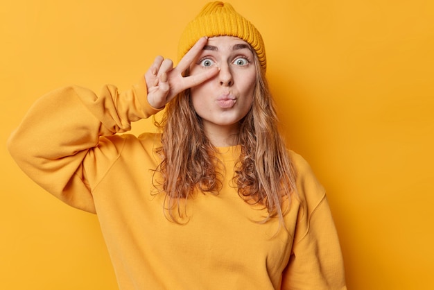 Wondered millennial girl with long hair keeps lips folded makes peace gesture over eye feels impressed dressed in casual clothes isolated on vivid yellow background enjoys life Body language concept