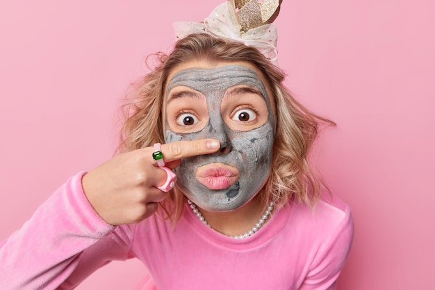 Free Photo wondered young european woman with hairstyle applies moisturizing clay mask on face touches nose has widely opened eyes wears crown jumper and necklace isolated over pink background beauty concept