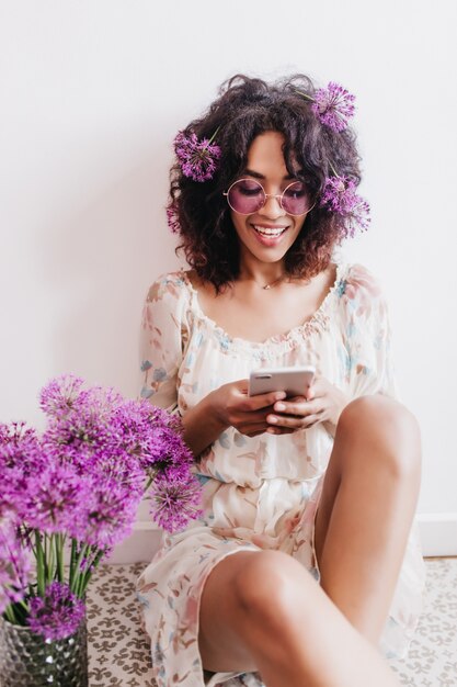 Wonderful black brunette woman texting message with smile. Indoor shot of pretty african girl sitting beside allium bouquet.