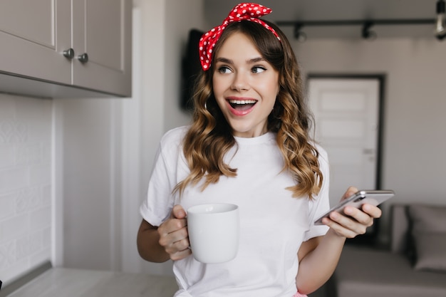 Wonderful caucasian girl holding phone and drinking coffee. Indoor portrait of pretty glad woman posing with smartphone and cup of tea.