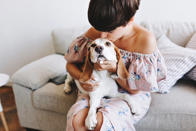 Wonderful girl with trendy short hairstyle kissing beagle dog while sitting on gray sofa