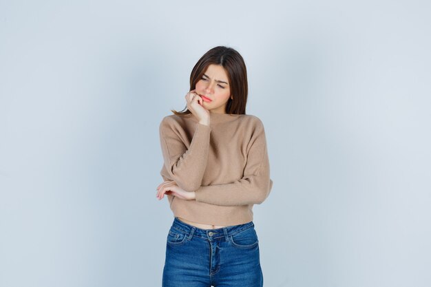 Wonderful lady in sweater, jeans keeping hand on chin while looking down and looking thoughtful , front view.