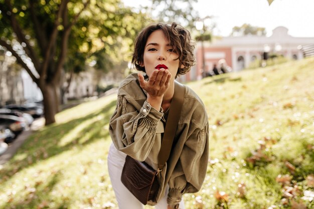 Wonderful woman in denim jacket and white pants blowing kiss outside. Brunette woman with red lips with handbag posing outdoors.