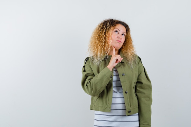 Wonderful woman in green jacket, shirt keeping finger under chin, looking up and looking thoughtful , front view.