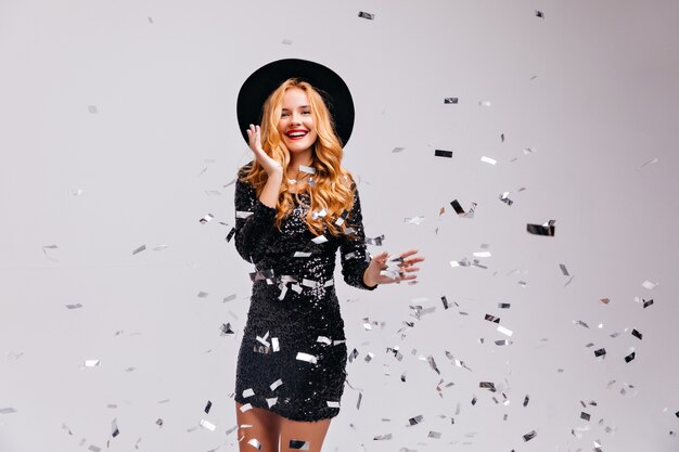 Wonderful young woman posing with confetti on white wall.  portrait of emotional girl in trendy hat expressing happiness.