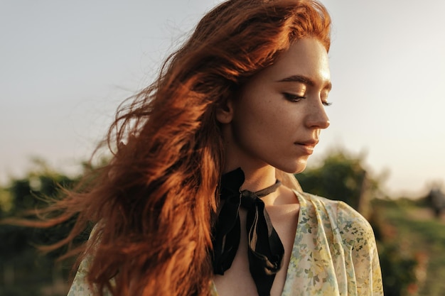 Wonderful young woman with wavy bright red hairstyle black bandage on neck and freckles in printed light clothes looking down outdoor