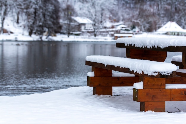 Wooden bench and a table near the lake surrounded by trees covered in the snow during the winter