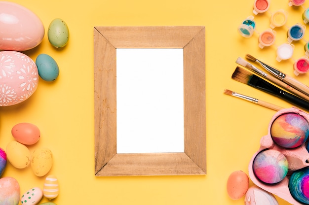 Free photo wooden blank white frame with easter eggs; paint brushes and watercolor paint on yellow background