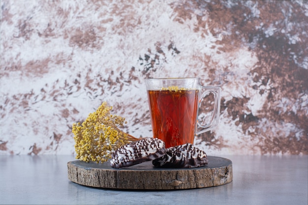 A wooden board of a glass cup of hot tea with cookies and mimosa flower