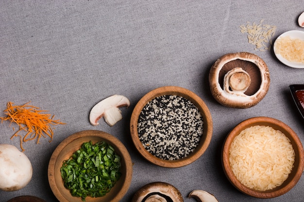 Wooden bowl of chives; sesame seeds; rice grains; mushroom and grated carrot on grey linen cloth