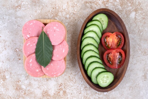 Wooden bowl of sliced sausage and chopped cucumbers on marble surface.