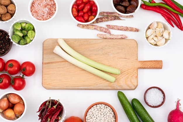 wooden cutting board with empty copy space surrounded by different vegetables in round bowl on white table