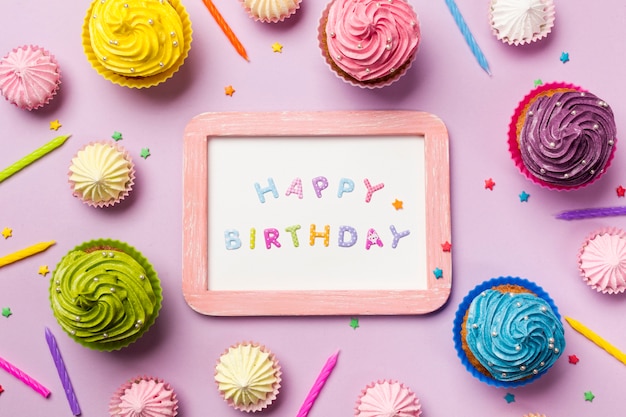 Free photo wooden happy birthday on white slate surrounded with muffins; aalaw; candles and sprinkles on pink background