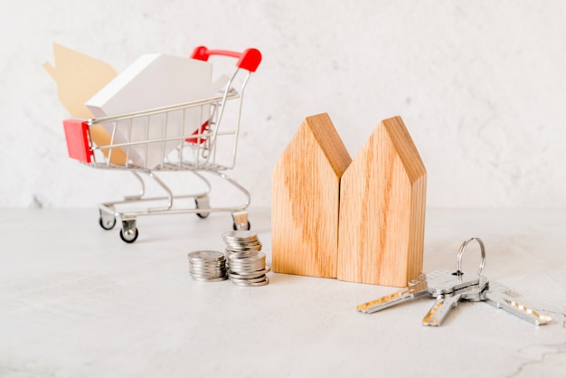 Free photo wooden house blocks; stack of coins; keys and small shopping cart on concrete backdrop