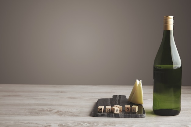 Wooden letter brick near sliced goat cheese and half empty green wine juice grape bottle isolated on marble stone board on white wooden table and gray neutral background