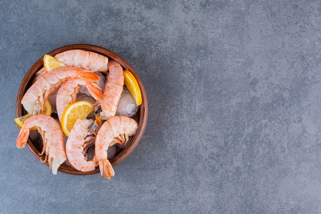 A wooden plate of delicious shrimps with ice cubes and sliced lemon on a stone surface