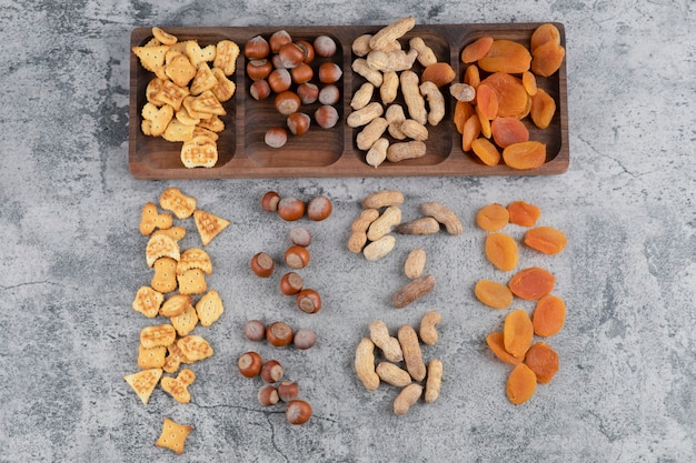 Free photo wooden plate full of nuts, crackers and dried apricots on marble surface.