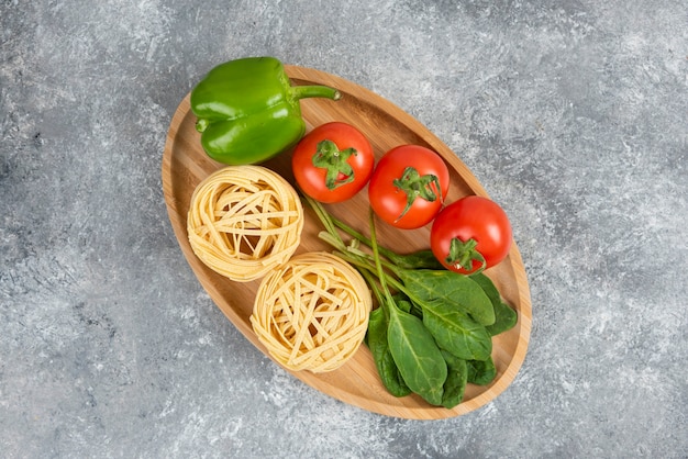 Wooden plate full of raw noodles and vegetables on marble surface.