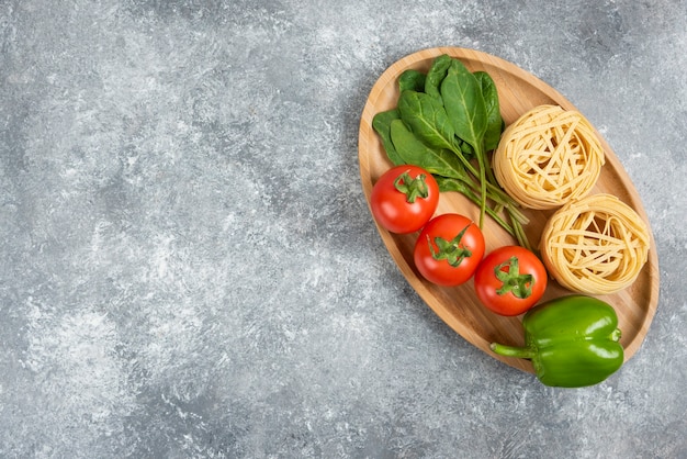 Wooden plate full of raw noodles and vegetables on marble surface.