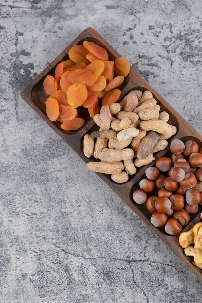 Wooden plate full of various nuts, crackers and dried apricots on marble surface.