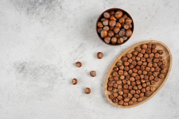 Wooden plate of hazelnut kernels and bowl of shelled hazelnuts on marble table. 