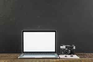 Free photo wooden table with blank screen laptop and retro photo camera on black background