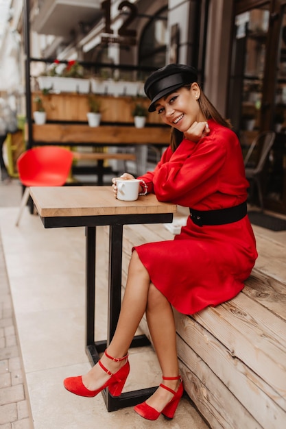 Free photo on wooden terrace near cafe young and beautiful girl dressed in bright red dress and red shoes drinks aromatic coffee and looks into camera