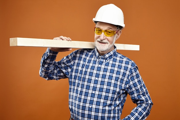 Free photo woodwork, craft and carpentry concept. confident positive senior mature woodman or carpenter with thick beard smiling , carrying wooden plank on his shoulder at blank orange wall