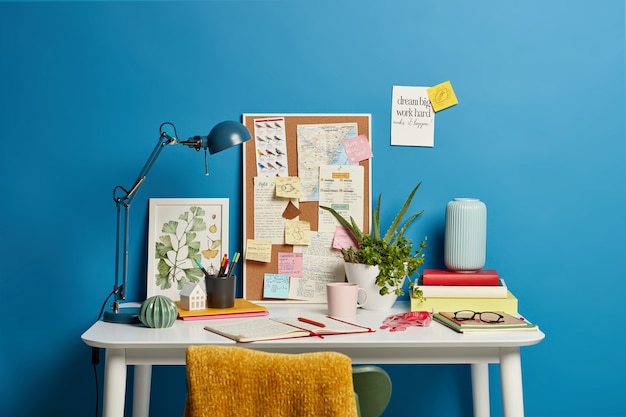 Free Photo work place of student. desktop with lamp, opened notebook, stationery and green indoor plant, mug of coffee sticky notes on blue. home office