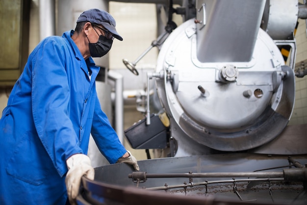 Worker in face mask controlling coffee roasting process. Coffee roaster working on roasting equipment. Man in mask and uniform Working with machinery appliance