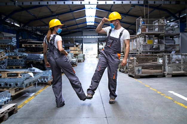 Workers wearing uniforms and hardhat at factory touching with legs and greeting due to corona virus and infection