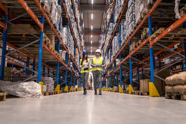 Workers with hardhats and reflective jackets walking through big warehouse aisle checking goods condition