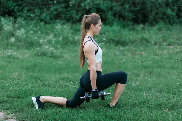 Working out outside. Pretty blonde athletic woman pumping up muscles with dumbbells 