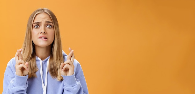 Free photo worried anxious hopeful young woman with fair hair in stylish oversized hoodie biting lower lip frowning concerned crossing fingers for good luck praying for dream come true against orange wall