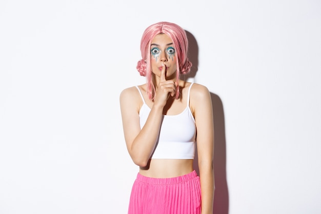 Worried attractive girl in pink anime hair and glitter, asking keep quiet, showing shush sign, press index finger to lips and looking concerned. Woman share a secret, standing.