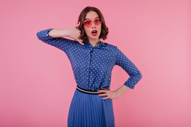 Worried beautiful young lady in blue skirt posing on pastel wall. Emotional lovely girl in sunglasses standing 