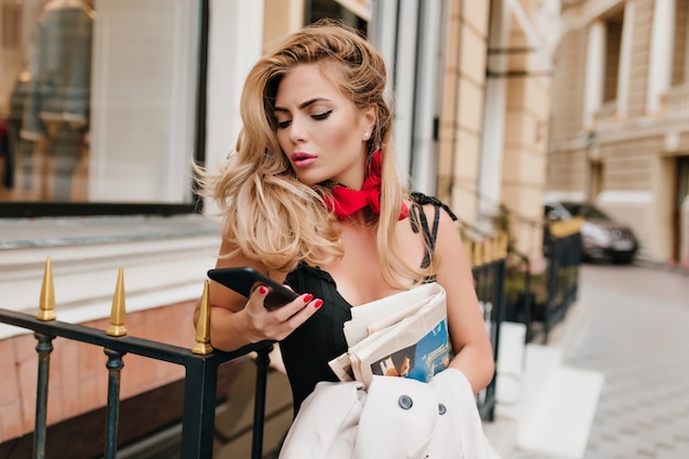 Worried blonde female model checking social networks while standing beside restaurant alone