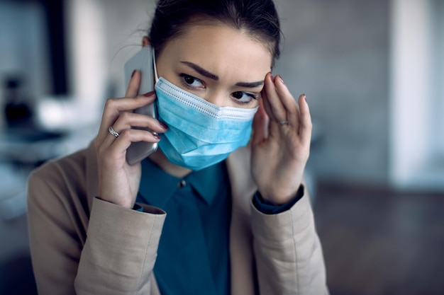 Worried businesswoman communicating over mobile phone while wearing protective face mask in the office