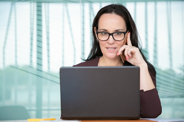 Worried businesswoman in glasses talking on mobile phone and making wide eyes, working at computer in office, using laptop at table