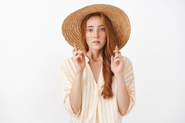 Worried concentrated good-looking ginger girl with freckles in cute summer straw hat and striped blouse crossing fingers for good luck looking concerned and worried praying, making wish