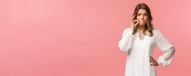 Worried cute blond girl give promise not tell anyone make zip lips gesture and frowning concerned heard someone secret keep silent about situation standing embarrassed pink background