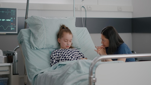 Free photo worried mother sitting beside little daughter praying while sleeping after illness surgery waiting for medical expertise in hospital ward. sick child wearing nasal tube during medicine treatment