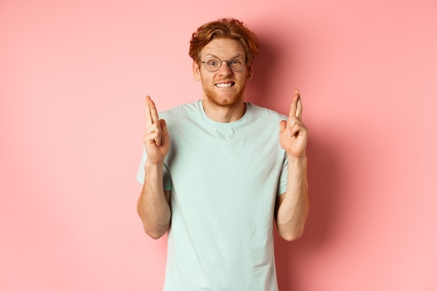 Worried redhead man waiting for results, expecting something with fingers crossed, biting finger and looking at something risky, standing over pink background.