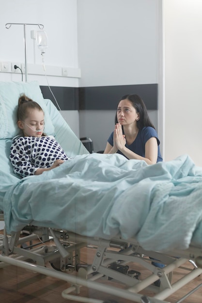 Free photo worried uneasy sad parent praying for sick hospitalized while in pediatric clinic ward room. unhappy attentive woman praying for ill daughter bad health condition in hospital patient room.