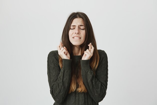 Worried young cute woman making wish with closed eyes and crossed fingers