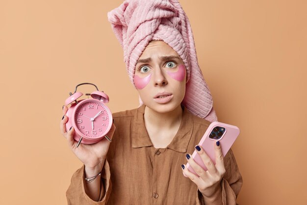 Worried young woman awakes late holds alarm clock and smartphone undergoes morning routines dressed in pajama wrapped towel on head isolated over brown background Awakening and rest concept