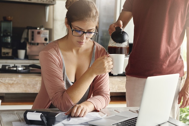 Worried young woman calculating family expenses and doing domestic budget using generic laptop and calculator in kitchen while her husband standing next to her and pouring hot coffee into her mug