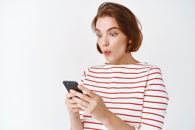 Wow check this out. Surprised and excited girl looking at mobile phone screen, online shopping with big discounts, standing in casual clothes against white wall