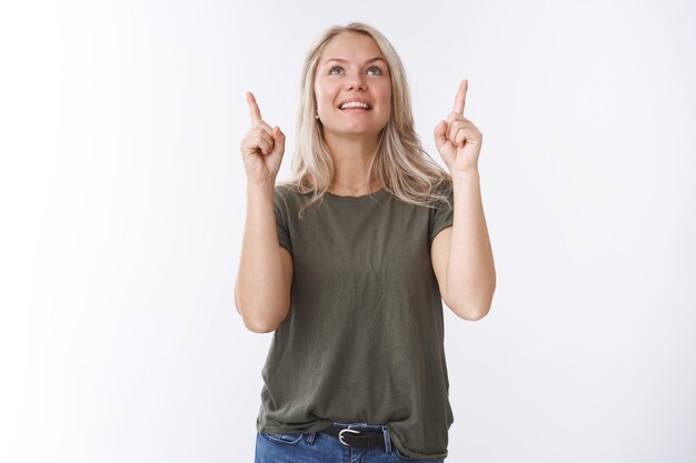 Wow how amusing. Portrait of fascinated amazed and joyful young beautiful woman with blond hair looking pointing up smiling from thrill and joy loving gazing sky over white background